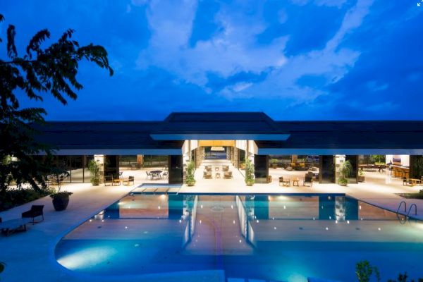 The image shows a luxurious, illuminated resort with an expansive pool and a modern building in the background under a twilight sky.