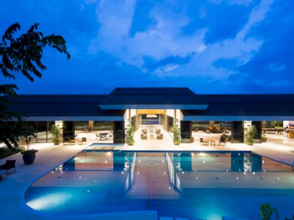 The image shows a luxurious, illuminated resort with an expansive pool and a modern building in the background under a twilight sky.