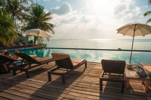 Poolside deck with lounge chairs, umbrellas, overlooking a serene infinity pool and ocean. Palm trees and blue sky complete the tranquil view.