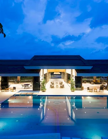 A modern building with a large illuminated swimming pool, lounge chairs, and open seating areas, set against a twilight sky.