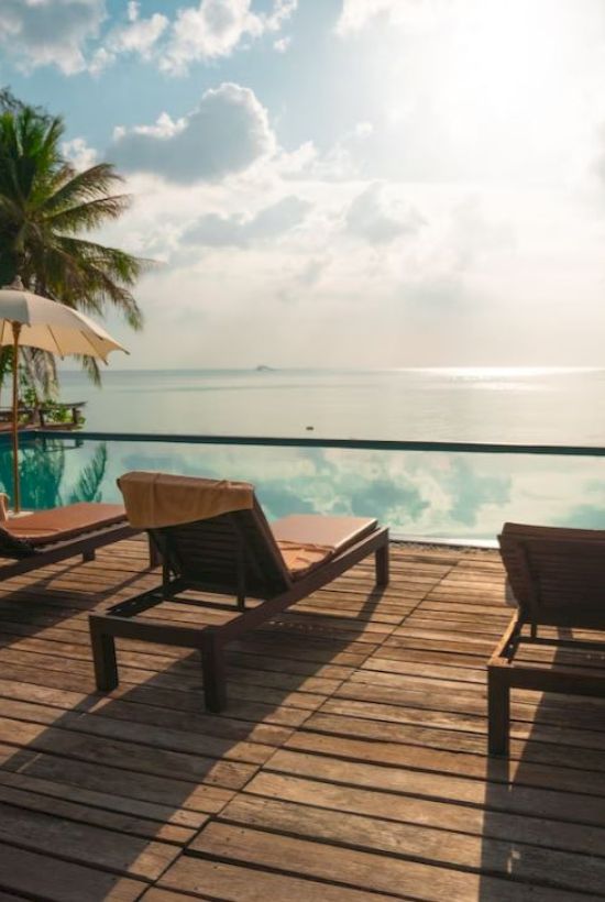 A serene poolside view with lounge chairs, palm trees, umbrellas, and a tranquil ocean backdrop under a partly cloudy sky.