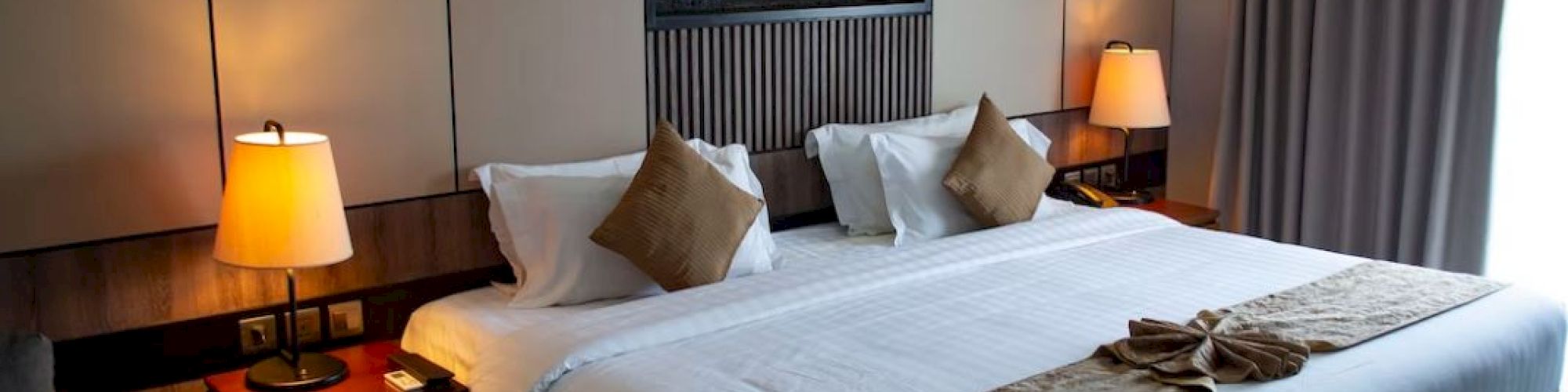 A cozy hotel room with a large bed, bedside tables with lamps, a decorative wall feature, and a window with curtains.
