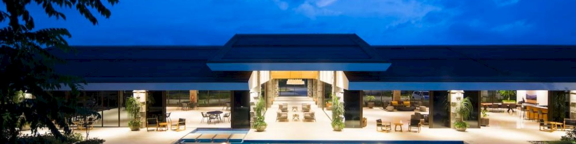 An illuminated luxury residence with a large swimming pool in front, set against a backdrop of a blue evening sky, light reflecting off the water.