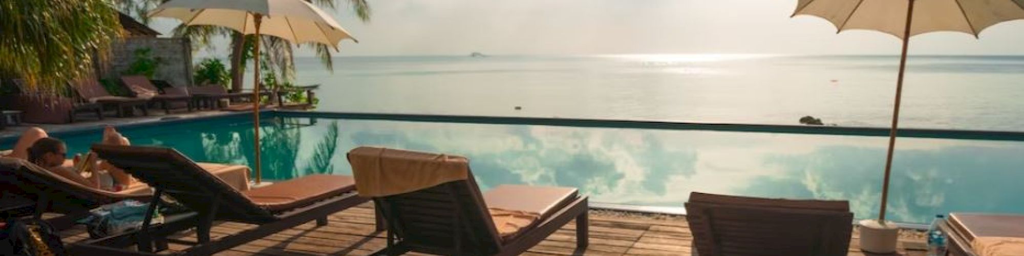 The image depicts a serene poolside area with lounge chairs and umbrellas under a sunny sky, overlooking a body of water, surrounded by palm trees.