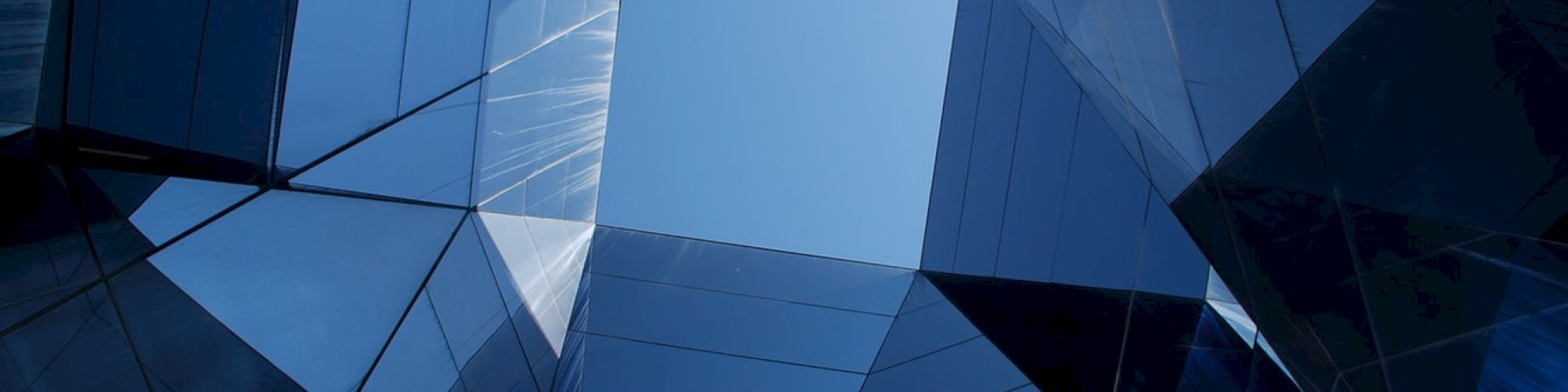 The image shows an abstract view of a skyscraper's mirrored glass walls, forming geometric patterns under a clear blue sky.