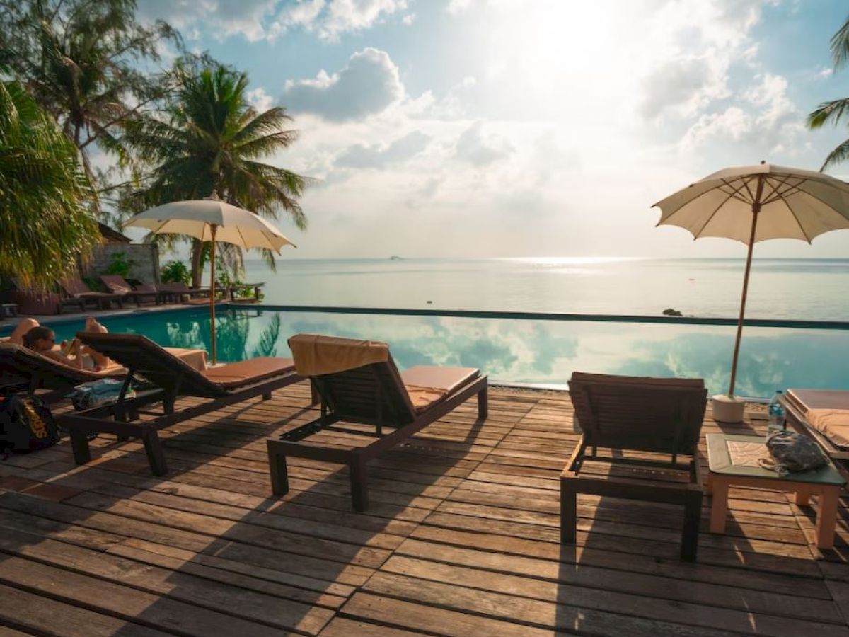 A serene tropical poolside scene with lounge chairs, umbrellas, and a view of the ocean under a bright sky with scattered clouds.