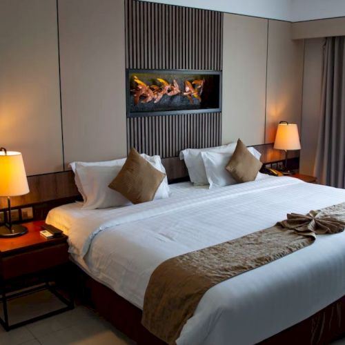 A well-lit hotel room features a large bed with pillows, bedside tables with lamps, and a window with curtains, with a wall decoration above the bed.