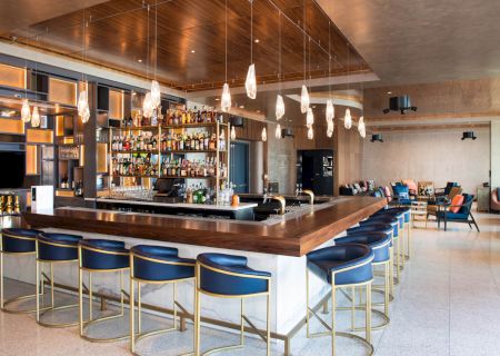 A modern bar with high seating, a stocked shelf of alcohol bottles, pendant lights, and a cozy seating area in the background, ending the sentence.