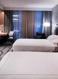 The image shows a modern hotel room with two neatly made beds, a flat-screen TV, a desk with a chair, and stylish decor.