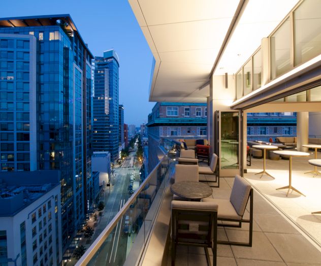 The image shows a modern balcony with seating, overlooking a cityscape with tall buildings and a street below during dusk.