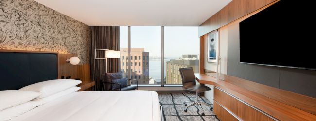 A modern hotel room features a large bed, wall-mounted TV, desk and chair, with a view of tall buildings through floor-to-ceiling windows.