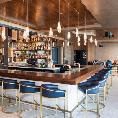 A modern bar with blue upholstered chairs, a variety of bottles on shelves, and pendant lights hanging above the counter in a spacious setting.