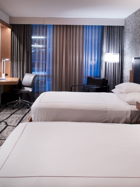 A modern hotel room features two neatly made beds, a wall-mounted TV, a desk with a chair, and stylish decor with a patterned accent wall.