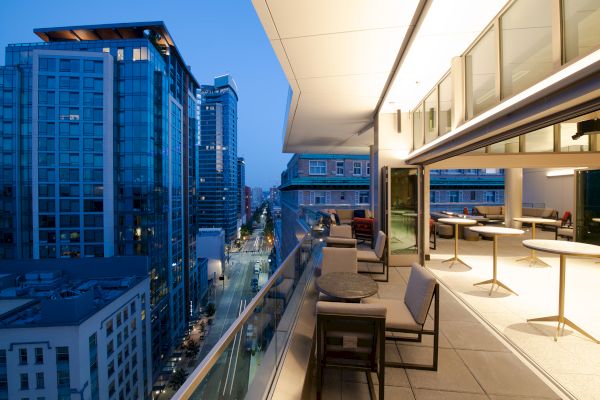 This image depicts a modern balcony or terrace at dusk in an urban setting, overlooking a street with tall buildings and city lights.