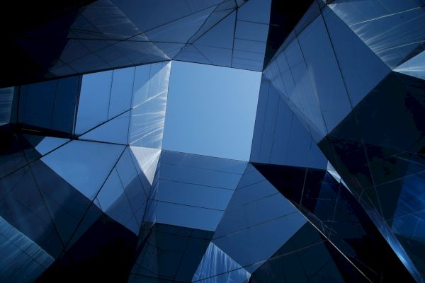 The image shows a geometric arrangement of reflective glass panels creating an open square space that reveals the sky above.