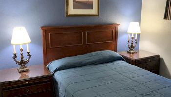 A bedroom with a wooden bed frame, blue bedding, two nightstands with lamps, and a framed picture on a blue wall. The floor is wooden.