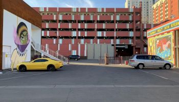 The image depicts a parking lot with a yellow car and a gray minivan, a colorful building facade, and a mural featuring a character with a purple scarf.