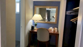 The image shows a small hotel room corner with a wooden desk, a chair, a lamp, and some coffee supplies, adjacent to a closet area with hangers.