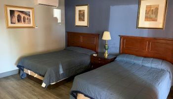 A hotel room with two double beds, topped with gray bedspreads, wooden headboards, framed art, wall-mounted AC, bedside lamp, and hardwood flooring.