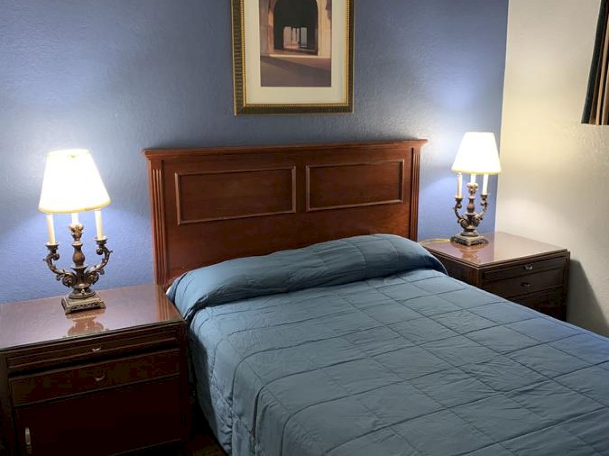 A cozy bedroom with a blue accent wall features a bed with a blue comforter, two bedside tables with lamps, and a framed picture above the headboard.