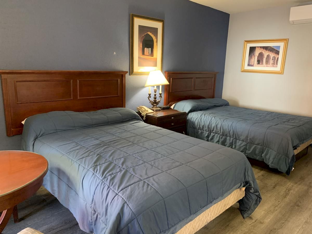 The image shows a hotel room with two double beds, blue bedding, a nightstand with a lamp, framed artwork, and part of a round table with chairs.