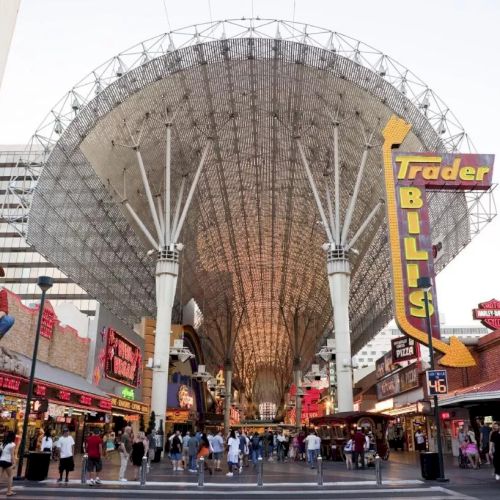 A busy street scene underneath a large canopy structure with numerous shops and neon signs, including one for "Trader Bills," and people walking around.