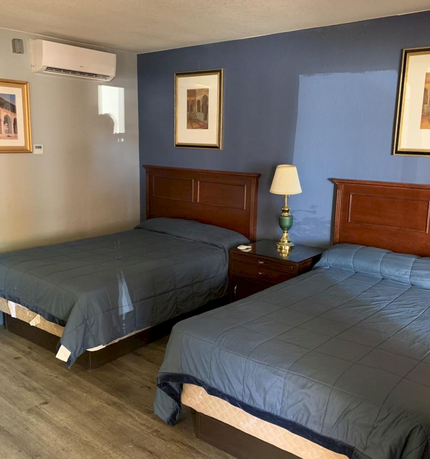 A room with two beds, a nightstand with a lamp, air conditioning, and framed art on the walls. The bedding is blue, and the floor is wood.