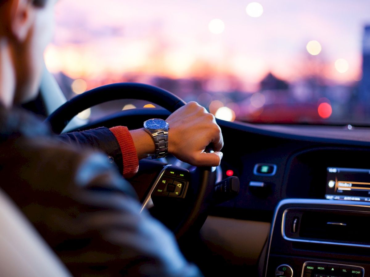 A person is driving a car at dusk, with a focus on their hand on the steering wheel and the car's dashboard, showing a blurred background.