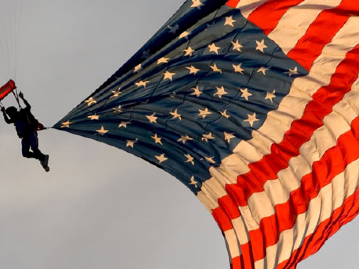 A person is parachuting with a large American flag (USA) trailing behind them in the sky. The flag is fully extended and prominently displayed.