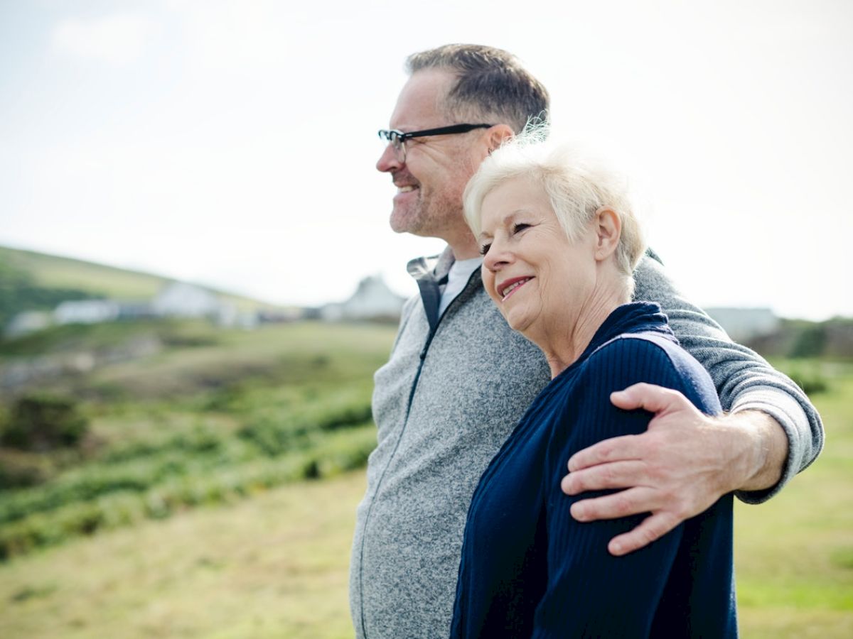 A smiling couple embraces while looking into the distance in a scenic outdoor setting with greenery and hills in the background.