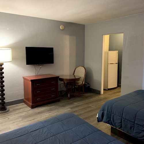 The image shows a hotel room with two beds, a flat-screen TV, a small table with a chair, a lamp, a mirror, and a partial view of a kitchen area.