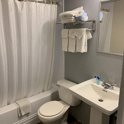 A bathroom with a shower curtain, toilet, pedestal sink, wall-mounted mirror, towel rack with towels, and a few toiletries on the sink.