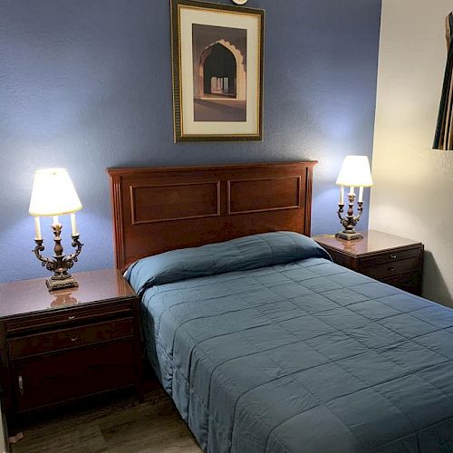 A bedroom with a bed, two nightstands, and table lamps against a blue wall with a framed picture above the headboard.