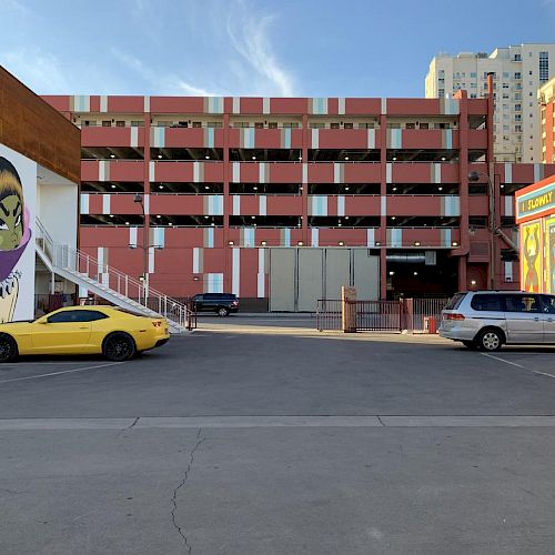 The image shows a parking lot with a yellow car and a silver minivan, a multi-story building, and murals on the walls, including a large face.