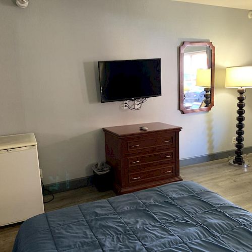 The image shows a room with a bed, a TV mounted on the wall, a dresser, a mirror, a lamp, and a small refrigerator.