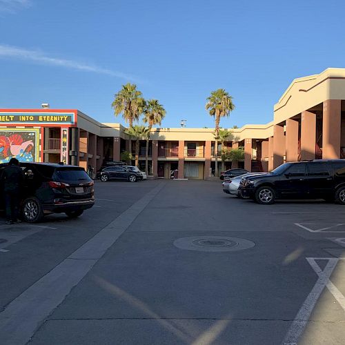 This image shows a parking lot outside a building complex with palm trees, parked cars, and a colorful mural on the left building.