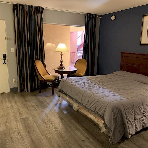 A hotel room with a bed, two chairs around a table with a lamp, wooden flooring, blue walls, and a framed picture above the headboard.