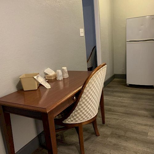 A small table with a container, tissues, small cups, and a chair, situated near a hallway leading to a refrigerator.