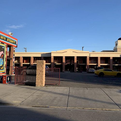 The image shows a building with a large mural on the left side and a parking lot with cars in front, under a clear blue sky.