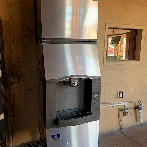 The image shows a commercial ice machine with a stainless steel exterior, placed against a wall next to a power outlet and some plumbing.