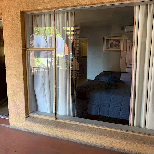 A view of a hotel room through a large window, with white curtains partially open, revealing a bed and some furniture inside.