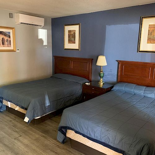 A hotel room with two double beds, blue and white bedspreads, framed art on the walls, a nightstand with a lamp, and wooden flooring.