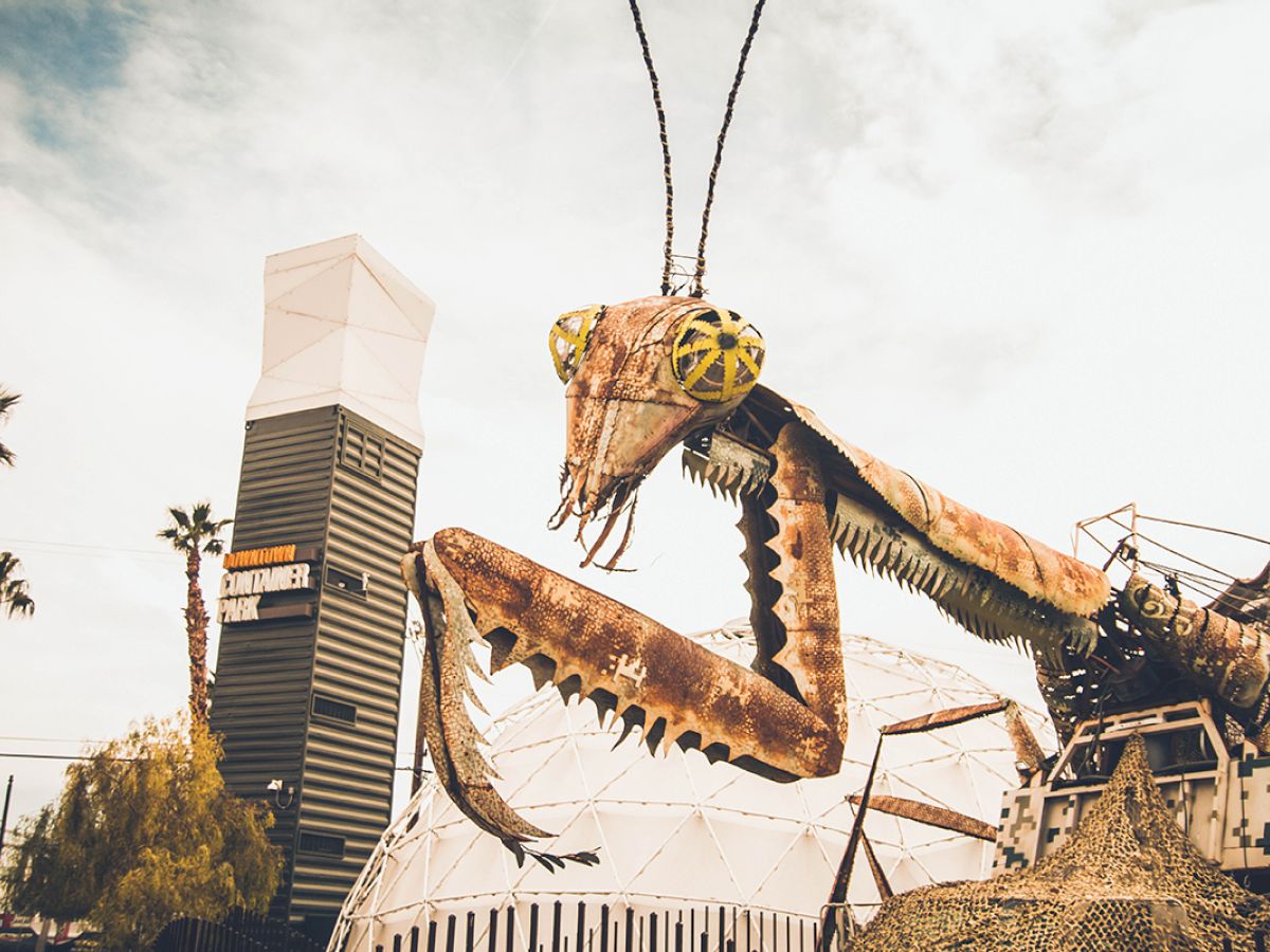 A giant mechanical praying mantis sculpture is displayed outdoors near a tall building and palm trees on a sunny day, creating an artistic scene.