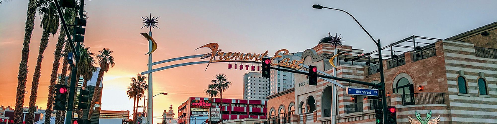 A city street scene at sunset with palm trees, a car, and buildings, including a prominent sign for "Fremont East District" in the background.