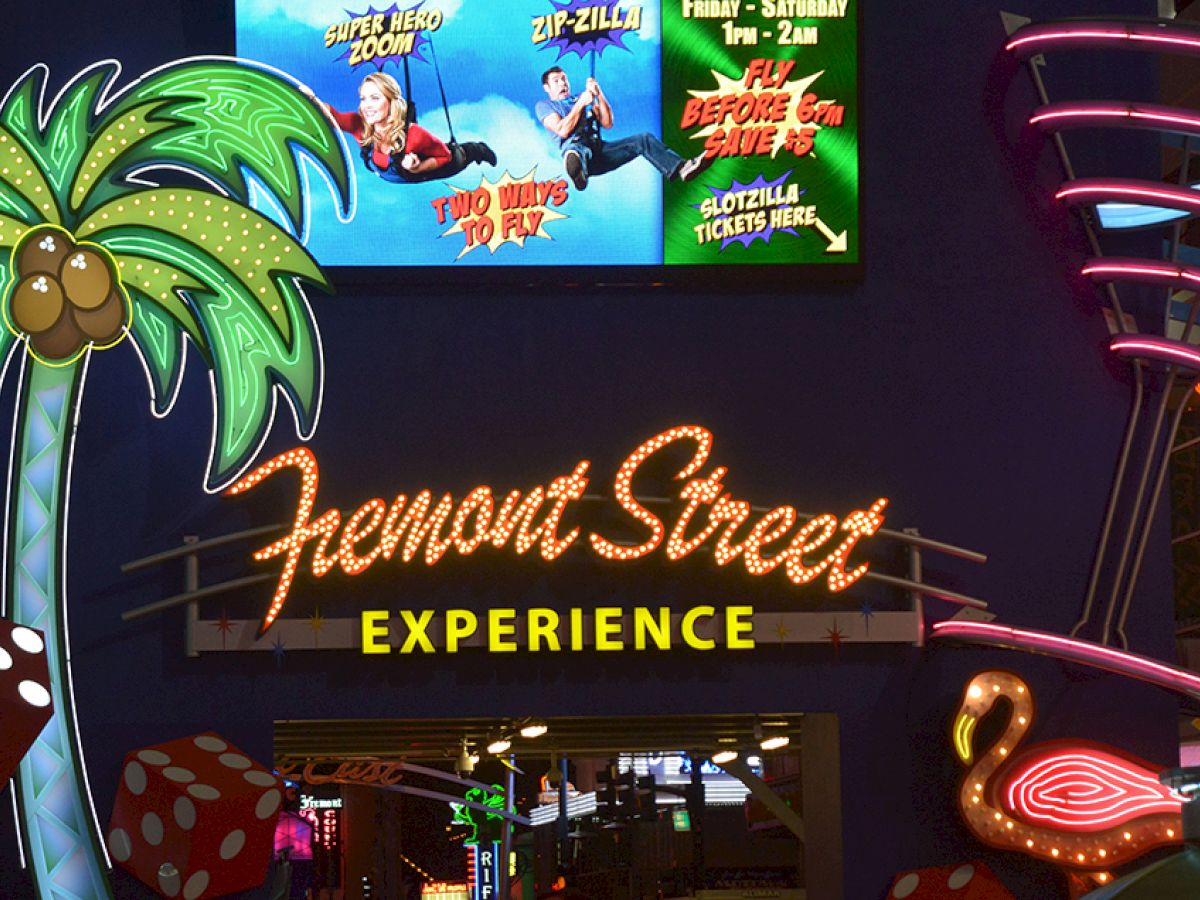 The image shows the illuminated entrance of Fremont Street Experience with neon signs including a palm tree, a flamingo, and a zip-line advertisement.