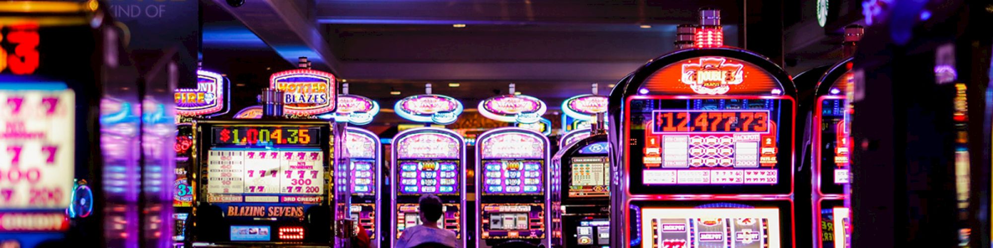 The image shows a dimly lit casino floor, filled with brightly illuminated slot machines and a couple of people seated, engaged in playing.