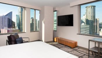 A modern bedroom with a city skyline view, featuring a bed, armchair, wall-mounted TV, wooden cabinet, and large windows showcasing tall buildings.