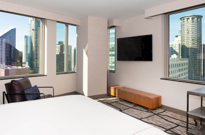 A modern bedroom with a city skyline view, featuring a bed, armchair, wall-mounted TV, wooden cabinet, and large windows showcasing tall buildings.