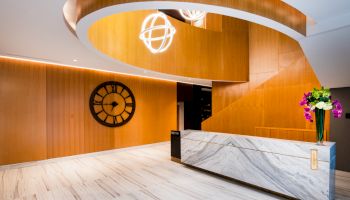 A modern lobby area with a white marble reception desk, wooden spiral staircase, large wall clock, and decorative lights and flowers.