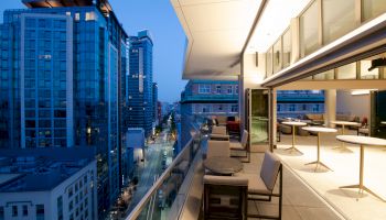 The image shows a modern cityscape view from a high-rise balcony with tables, chairs, and buildings at dusk, illuminated by lights along the street below.
